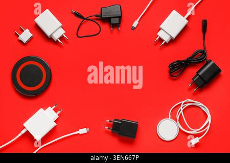 Modern electronic device chargers and adapters for mobile technology on vibrant red background, various white and black charging cables, wall plugs Stock Photo