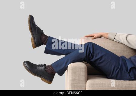 Relaxed businessman in blue suit and black shoes with patterned socks resting on a beige sofa, isolated on gray background Stock Photo