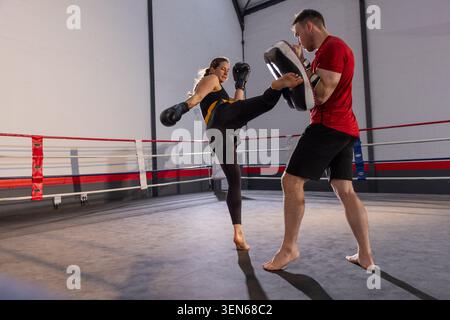 Athlete wearing black leggings yellow top kicking with gloves, trainer holding focus pads in ring Stock Photo