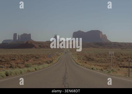 Long empty road leading through a dry desert toward large red rock mesas under a clear blue sky, capturing a classic american southwest travel scene. Stock Photo