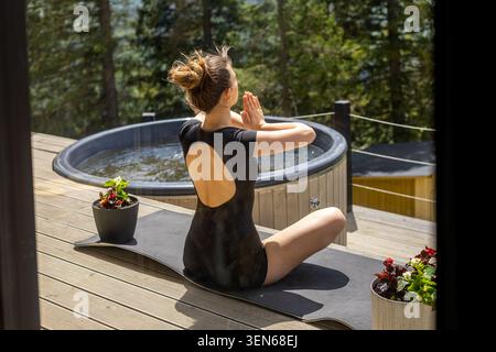Woman Meditating On Outdoor Spa Terrace. Woman practicing seated yoga meditation on a wooden terrace beside a hot tub, surrounded by forest and Stock Photo