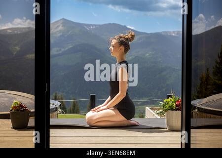 Woman Meditating On Terrace With Mountain View. Woman sitting in meditation on a yoga mat, overlooking peaceful mountains from a sunny wooden terrace. Stock Photo