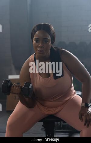African American woman doing bicep curl at gym bench holding black hex dumbbell pink tank top Stock Photo