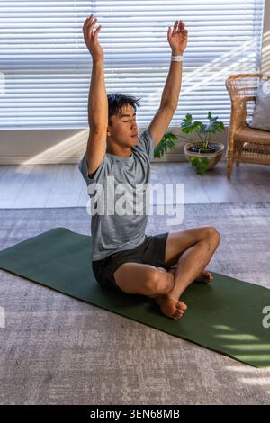 Asian man practicing seated yoga in living room on green mat, wearing white wristwatch Stock Photo