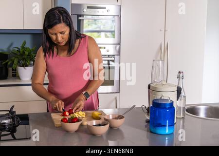 Mature woman in pink top using knife slicing strawberries, placing banana pieces at kitchen island Stock Photo