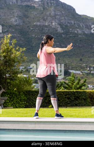 Asian woman balancing on stone pool edge in backyard garden, wearing sleeveless pink top Stock Photo