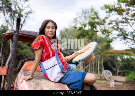 Woman farmer straw hat tractor apron red shirt garden rural outdoor portrait, woman farmer holding straw hat sitting on tractor in rural garden with a Stock Photo