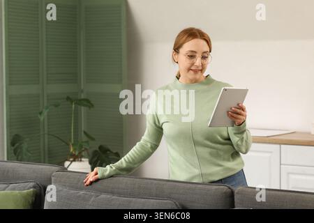 Mature woman using digital tablet for online learning and remote work in modern home living room, she is wearing glasses and a light green sweater, ho Stock Photo