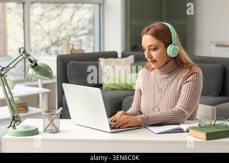 Mature woman in headphones working on laptop at home office desk, remote work and online learning concept, the woman is focused on her computer Stock Photo