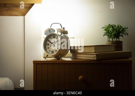 Vintage alarm clock and stack of books on wooden nightstand with plant, cozy bedroom interior, a small green plant in a woven pot adds a touch of natu Stock Photo