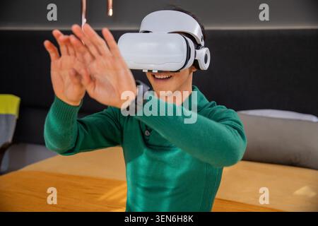 Man is wearing white VR headset and headphones and reaching over wooden table in office lounge Stock Photo