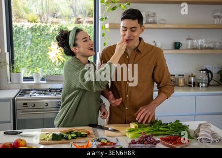 Couple laughing and feeding while preparing cucumbers, cherry tomatoes, knives on kitchen island Stock Photo