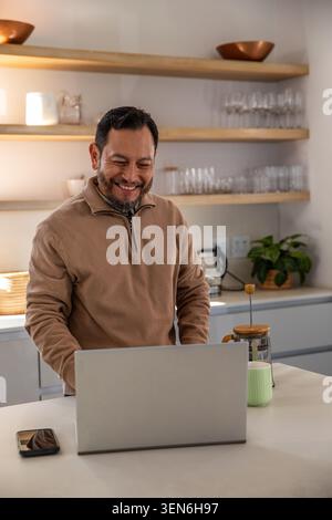 Mature Asian man standing at kitchen island using laptop and smiling with phone and French press Stock Photo