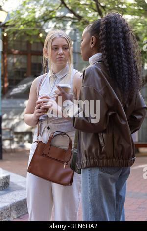 Diverse female friends chatting while holding takeaway coffee cups in tree-lined urban courtyard Stock Photo