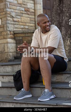 Man sitting on stone steps in front of masonry building, smiling and gesturing with backpack Stock Photo