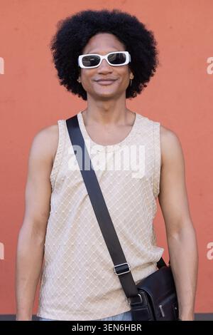 African American man standing against red wall, wearing beige top, white sunglasses, crossbody bag Stock Photo