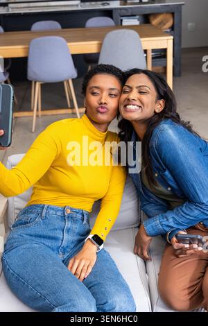 Indian and African American female friends taking selfie with smartphone, second phone on sofa Stock Photo