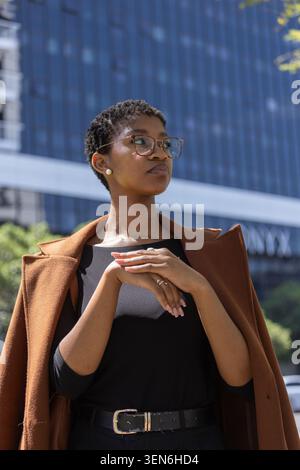 African woman standing in front of glass office building, brown coat draped, hands clasped, glasses Stock Photo