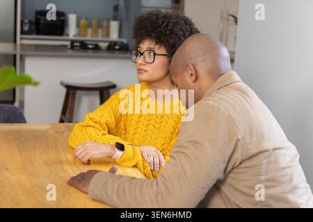 Couple sitting at wood table in home kitchen, woman wearing yellow sweater checking smartwatch Stock Photo