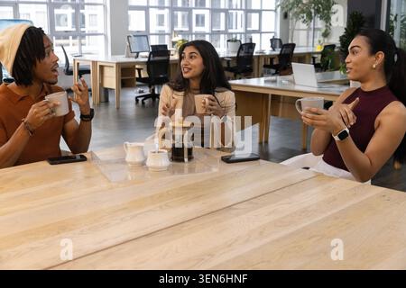 Diverse coworkers sitting at long table in office holding mugs and French press, smartphones nearby Stock Photo