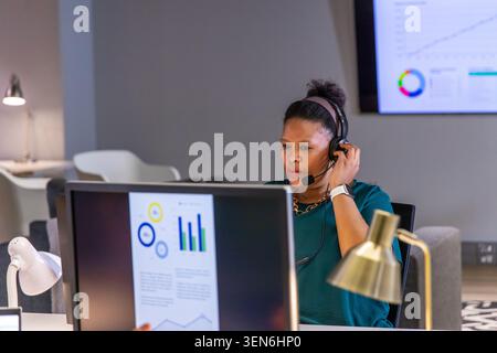African American woman analyzing charts on dual monitors at desk, wearing teal top and headset Stock Photo