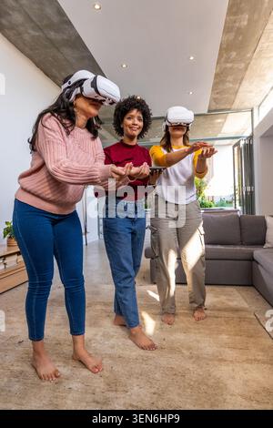 Diverse female friends using white VR headsets and holding tablet, standing barefoot in living room Stock Photo