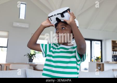 African American child lifting white VR headset behind gray couch at home in green-striped shirt Stock Photo