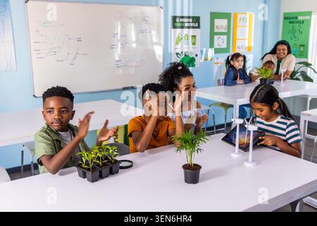 Diverse children clapping and using tablet to study mini wind turbines and plants in classroom Stock Photo