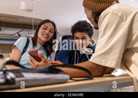 Diverse teenage classmates leaning over smartphone at campus lounge, backpacks and headphones. Students, collaboration, study, technology, university, Stock Photo