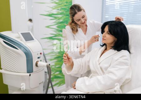 Smiling female cosmetologist consulting with middle-aged woman client in modern salon, examining reflection in hand mirror after facial treatment Stock Photo