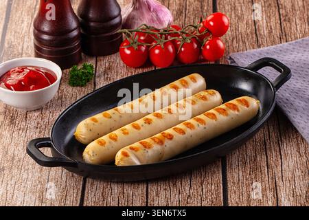 Three golden grilled sausages served in black cast iron skillet on rustic wooden table with cherry tomatoes and ketchup side dish Stock Photo