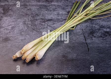 Fresh lemongrass stalks arranged diagonally on textured dark gray stone surface. Natural lighting highlights pale green and yellow hues. Ideal for cul Stock Photo