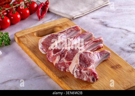 Four raw lamb chops arranged on a wooden cutting board, surrounded by bright red cherry tomatoes, dried red chili peppers, fresh green parsley on a li Stock Photo