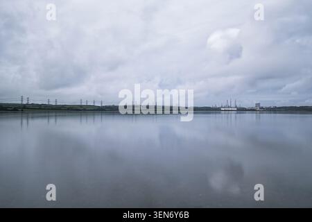 A grey, overcast sky reflected in the calm, tidal waters of the Pembroke River. On the horizon is Pembroke Refinery and Power Station. Stock Photo