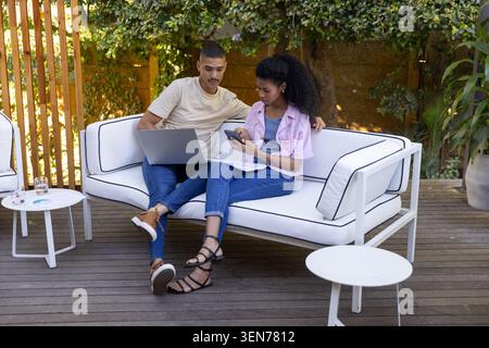 African American couple sitting on outdoor sofa on deck, using laptop and holding phone and papers Stock Photo