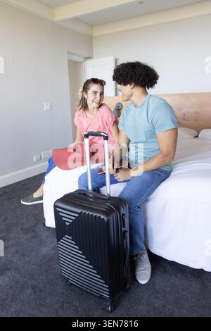 Couple sitting in hotel room, holding passport and phone with red sun hat beside black suitcase Stock Photo