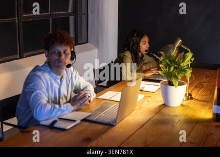 Diverse coworkers in headsets making calls while typing on laptops at wooden desk with notebooks Stock Photo
