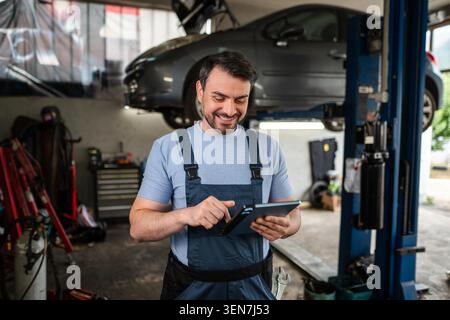Smiling mechanic using a tablet in a workshop with a lifted car Stock Photo