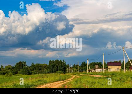 Massive dramatic cumulus clouds tower over a peaceful rural path, casting dynamic shadows across the lush green meadows and small countryside village Stock Photo