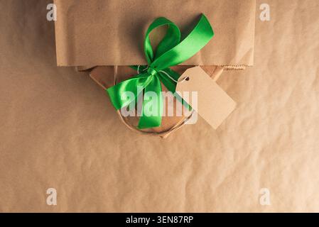 Gift box made from craft paper, tied with a bright green satin ribbon and a blank tag, peeking out from a brown paper shopping bag against a kraft bac Stock Photo