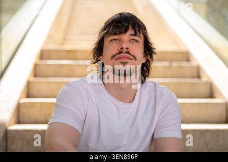 Young 25 year old man sits on steps and looks into the distance in an urban environment Stock Photo