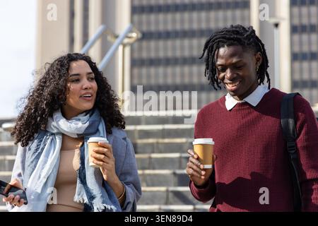 Diverse coworkers walking on office steps, wearing sweater and coat, holding coffee cups and phone Stock Photo