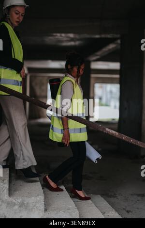 Two female engineers in high-visibility vests inspecting an unfinished building while descending concrete stairs Stock Photo