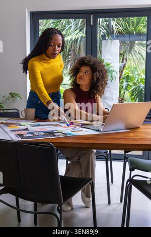 African American female team reviewing prints and pointing at layouts near laptop at table Stock Photo