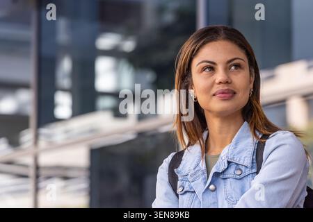 African American woman gazing right near glass facade in denim jacket backpack, copy space Stock Photo