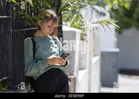 Adult female leaning against black fence on path, using phone, wearing sweater and black backpack Stock Photo