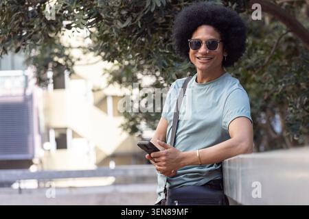 African American man in green tee leaning on railing at plaza, holding phone smiling, copy space Stock Photo