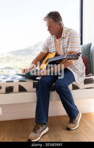 Man sitting on built-in window bench with acoustic guitar across lap, writing notebook with pen Stock Photo