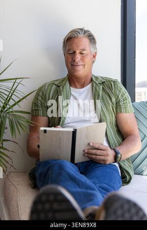 Senior man sitting on cushioned bench beside tall window holding open notebook, wearing green shirt Stock Photo