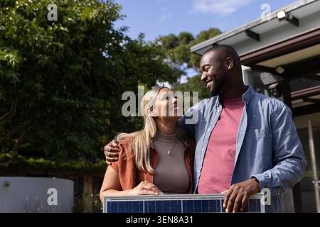Couple holding rectangular solar panel at suburban home porch under mature trees, smiling warmly Stock Photo